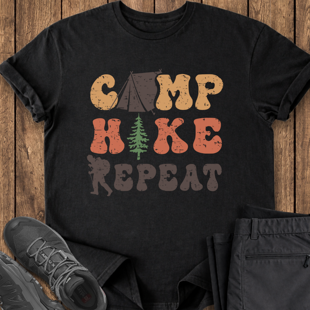 Black t-shirt with 'Camp Hike Repeat' design on a wooden surface with shoes and pants.