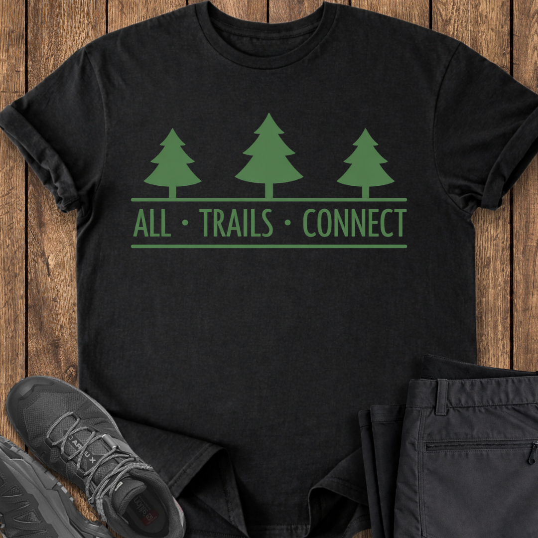 Black t-shirt with green tree design and 'All Trails Connect' text on a wooden surface with shoes and pants.