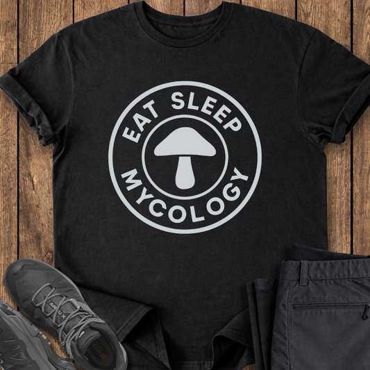Black t-shirt with 'Eat Sleep Mycology' design on a wooden surface with shoes and pants. mushroom hunting foraging