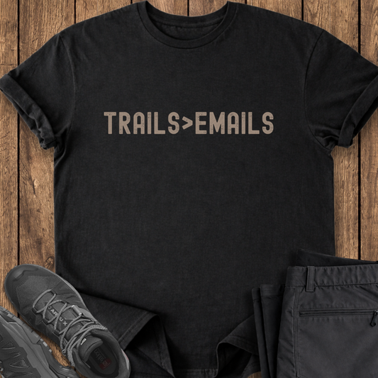 Black t-shirt with 'TRAILS>EMAILS' text on a wooden surface with shoes and pants.