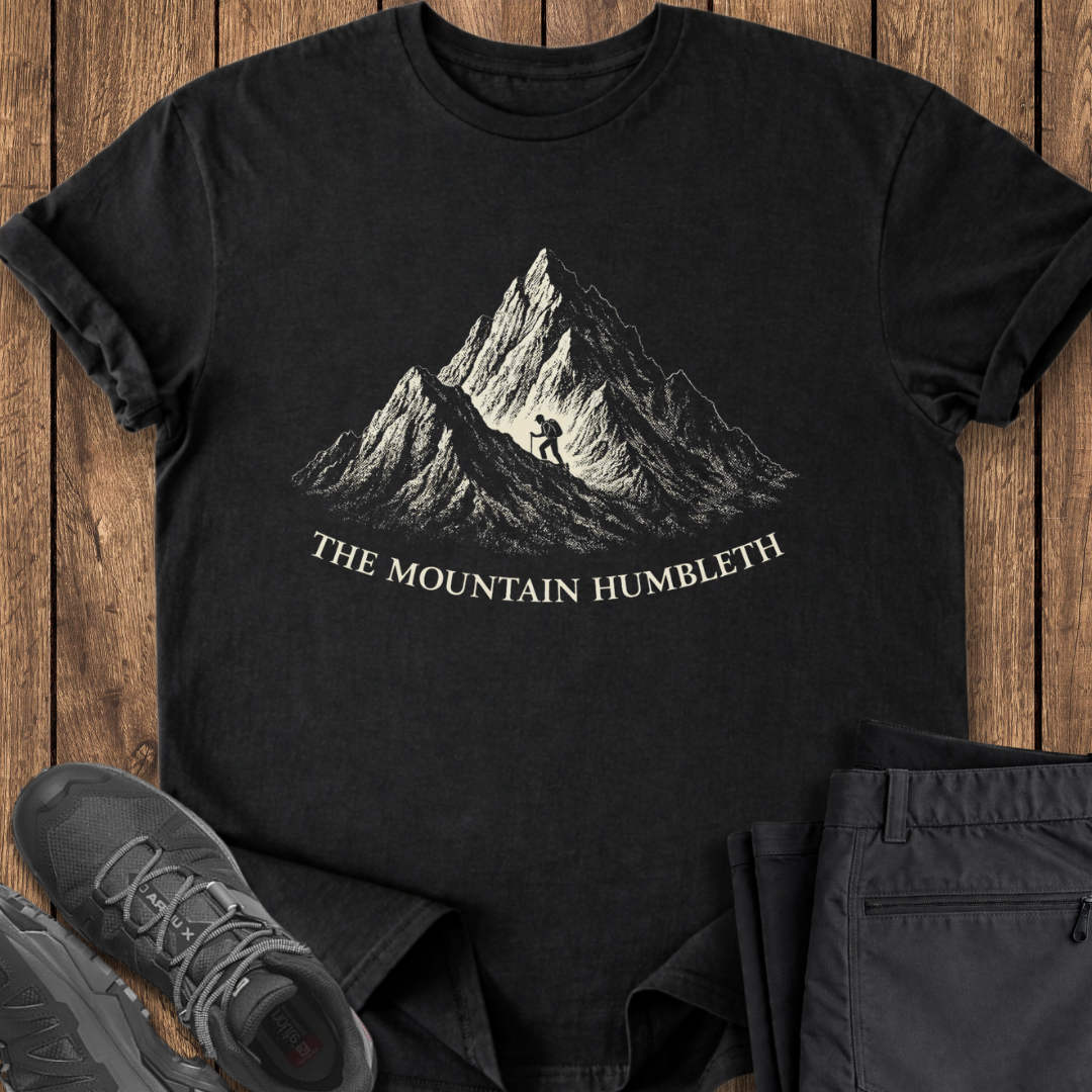 Black t-shirt with mountain graphic and text on a wooden surface with shoes and pants. funny mountain adventure t shirt