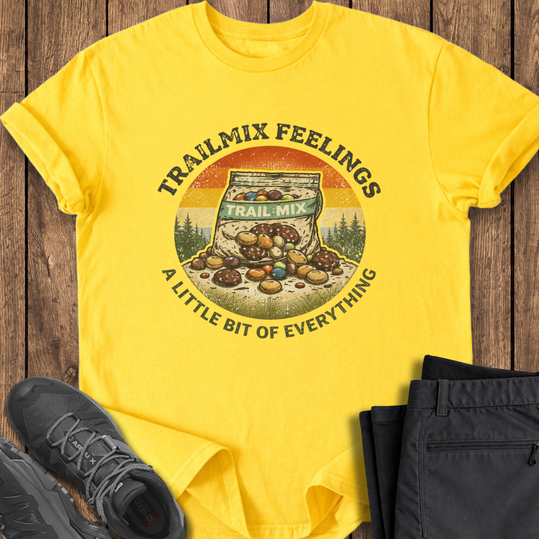 Yellow t-shirt with 'Trailmix Feelings' graphic on a wooden surface with shoes and pants. funny hiking t shirt