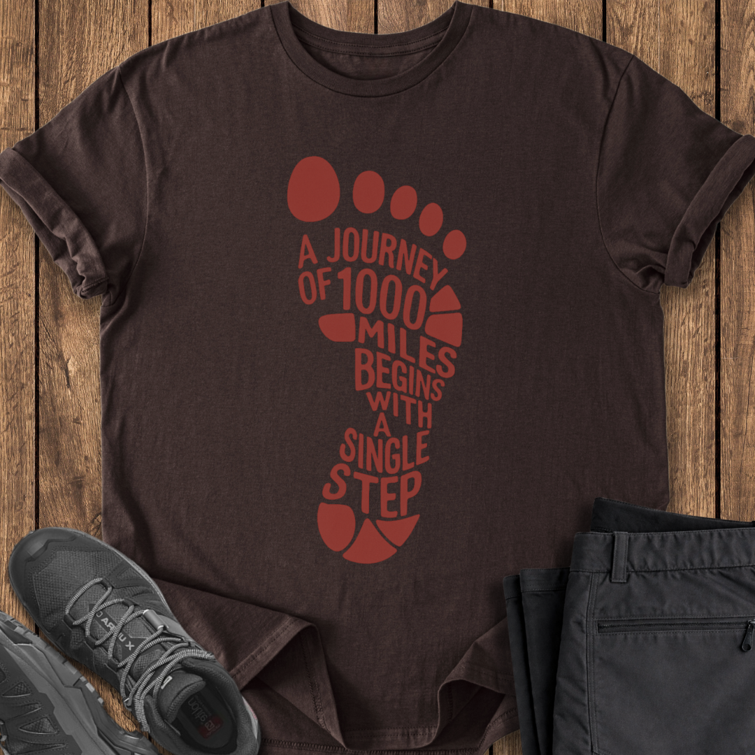 Brown t-shirt with red footprint design and text on a wooden surface, accompanied by black shoes and pants.