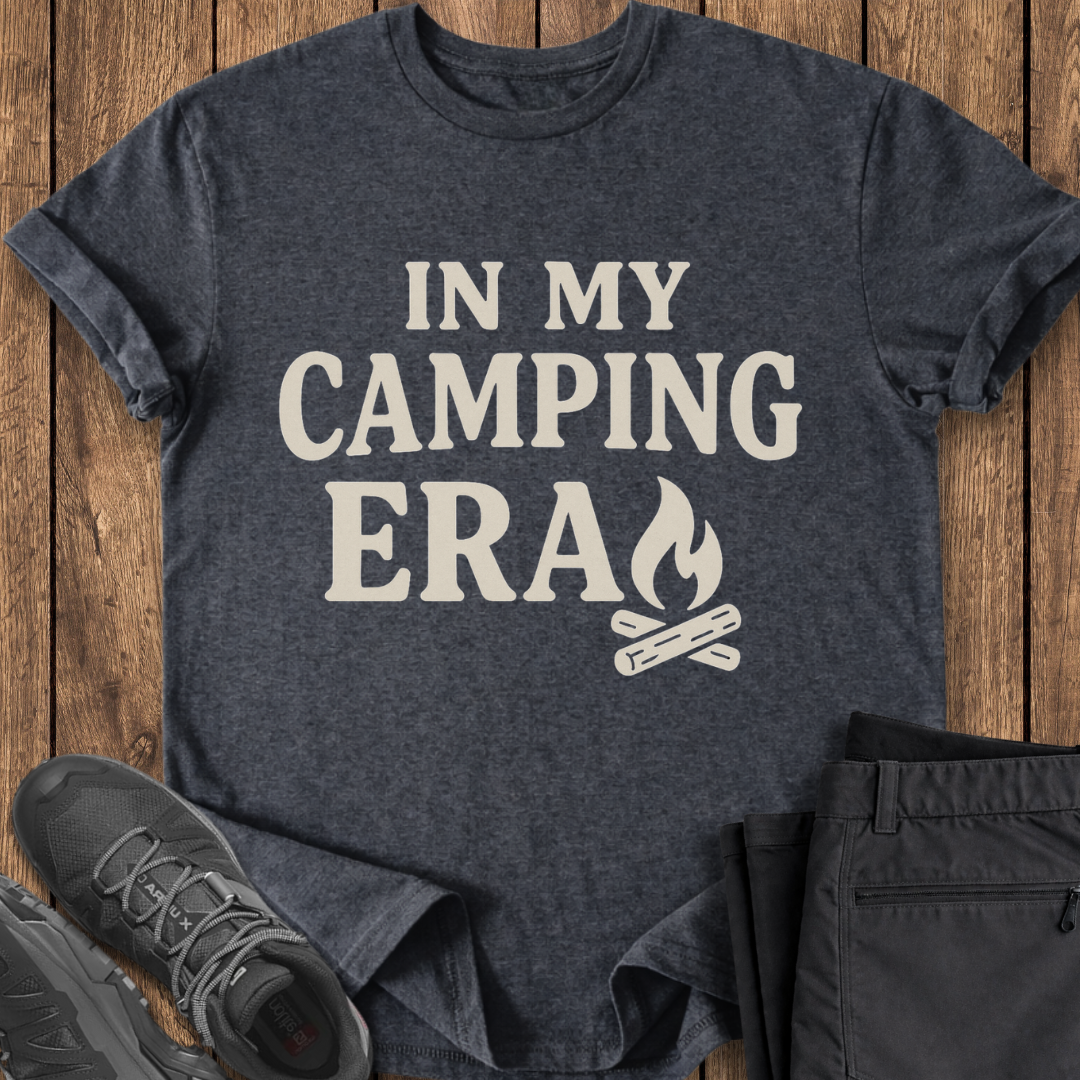 Dark gray t-shirt with 'In My Camping Era' text and campfire graphic on a wooden surface with shoes and pants. camp tshirt