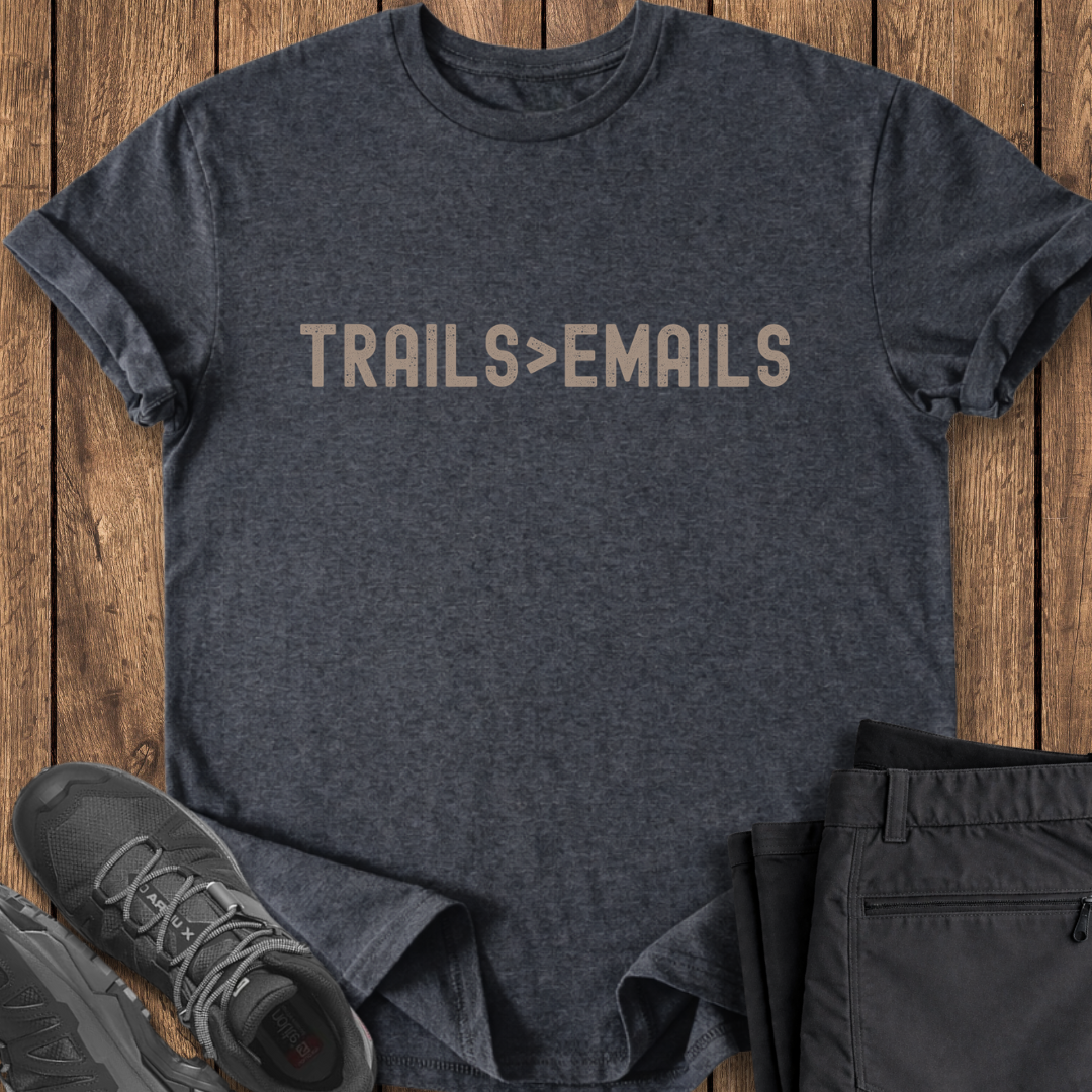 Dark gray t-shirt with 'TRAILS > EMAILS' text on a wooden surface, with shoes and pants in the foreground.