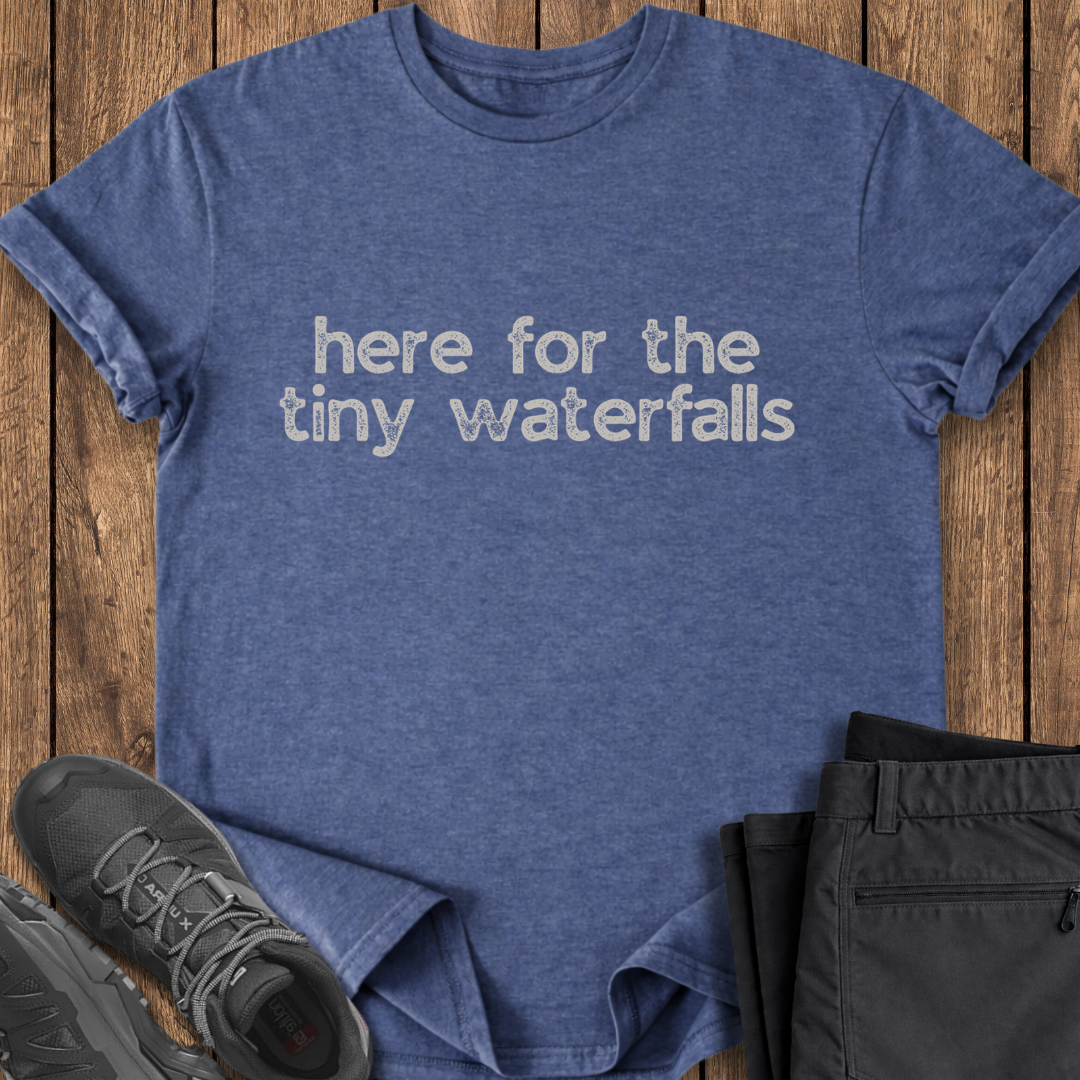 Blue t-shirt with 'here for the tiny waterfalls' text on a wooden surface with shoes and pants. hiking t shirt
