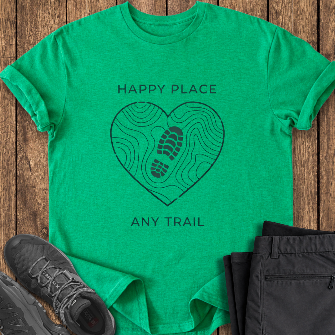 Green t-shirt with 'Happy Place, Any Trail' design on a wooden surface with shoes and pants. mindfulness hiking t shirts