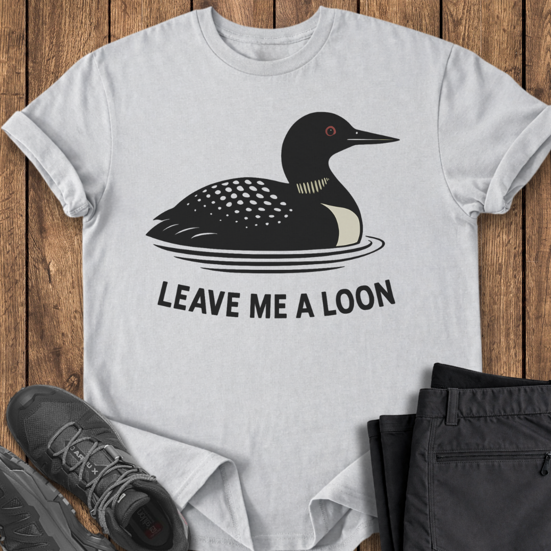 White t-shirt with a loon graphic and text 'Leave Me A Loon' on a wooden surface.