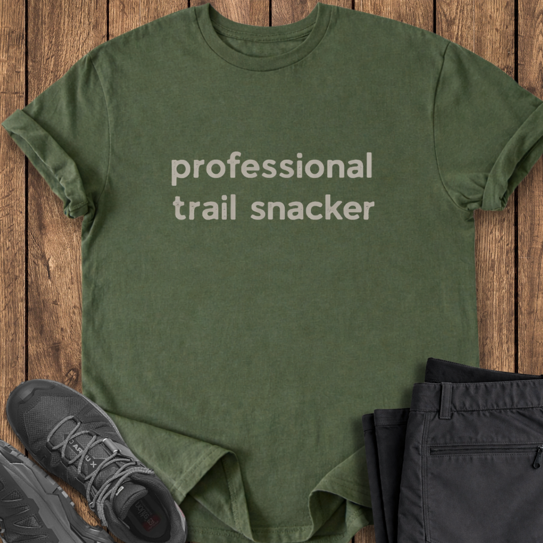 Green t-shirt with 'professional trail snacker' text on a wooden surface with shoes and pants. funny hike t shirt