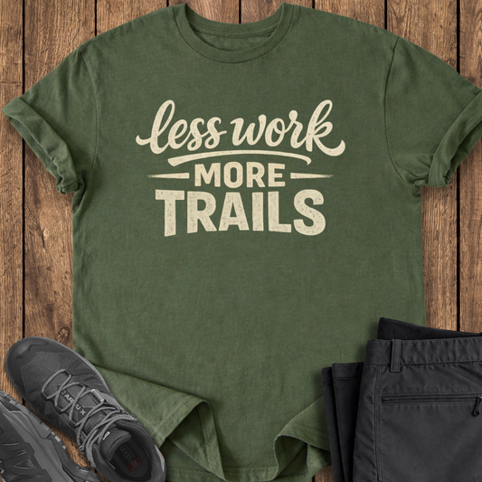 Green t-shirt with 'less work more trails' text on a wooden surface with black pants and shoes.