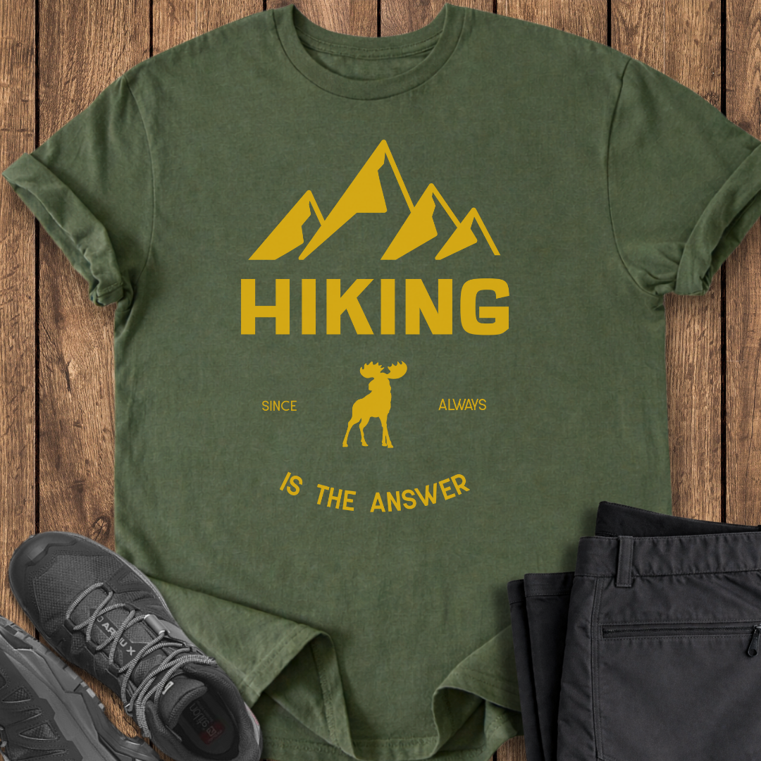 Green t-shirt with yellow mountain and moose design on a wooden surface with black pants and shoes. hiking mental health t shirt