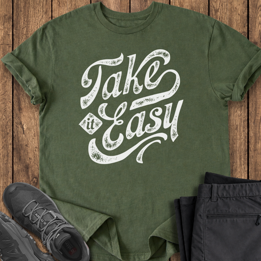 Green t-shirt with 'Take It Easy' text on a wooden surface with shoes and pants.