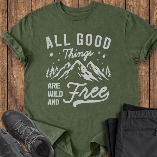 Green t-shirt with mountain graphic and text on a wooden surface with shoes and pants.