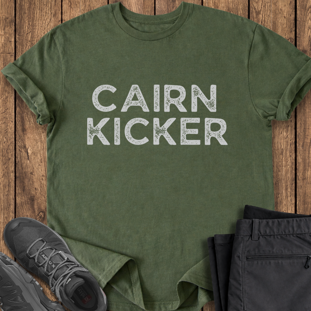 Green t-shirt with 'CAIRN KICKER' text on a wooden surface with shoes and pants. hiking t shirt