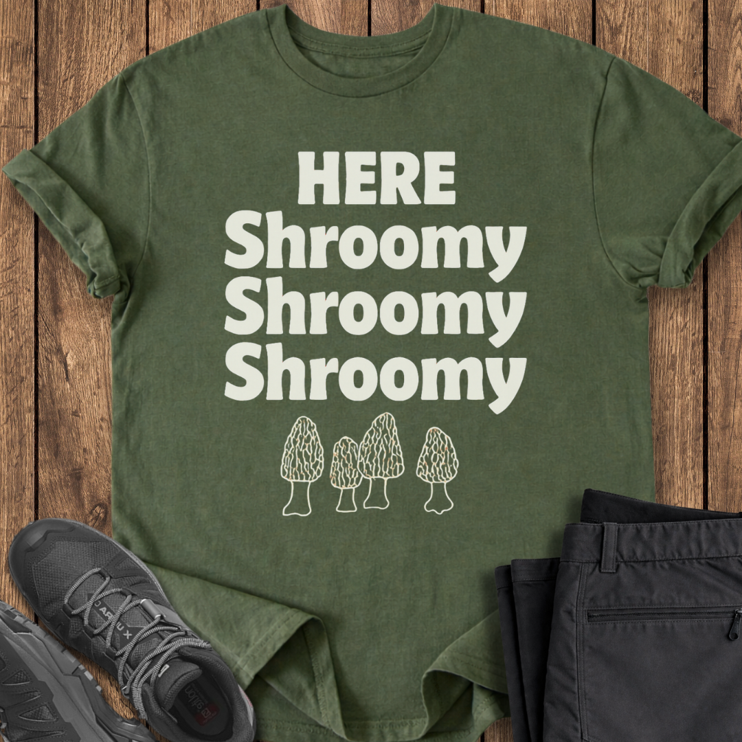 Green t-shirt with 'HERE Shroomy Shroomy Shroomy' text and mushroom graphics on a wooden surface, with shoes and pants for scale.
