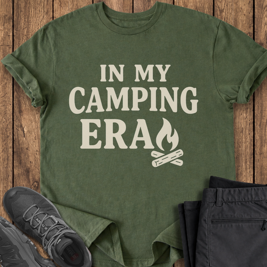Green t-shirt with 'In My Camping Era' text and campfire graphic on a wooden surface with shoes and pants. camper camp t shirt