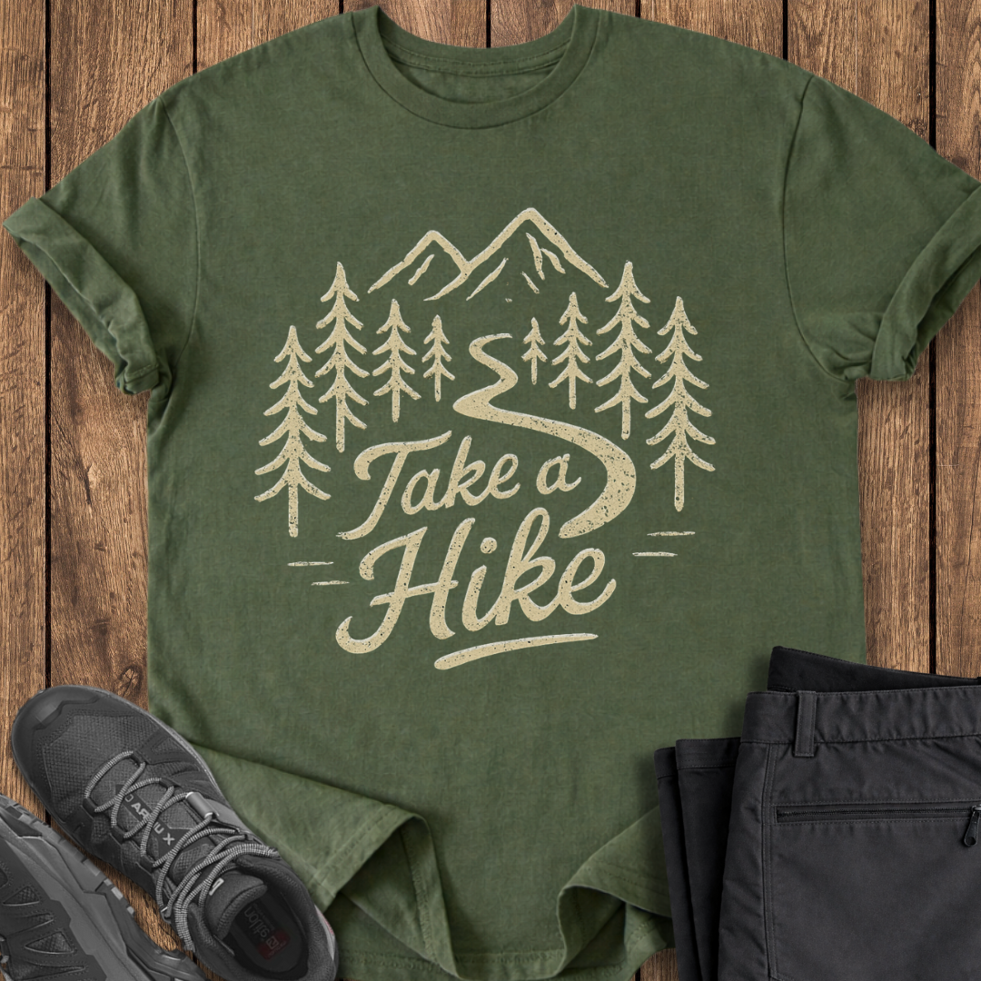 Green t-shirt with 'Take a Hike' design on a wooden surface with hiking shoes and pants.