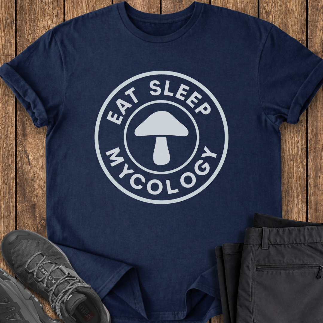 Navy blue t-shirt with 'Eat Sleep Mycology' design on a wooden surface with shoes and pants. mushroom hunting