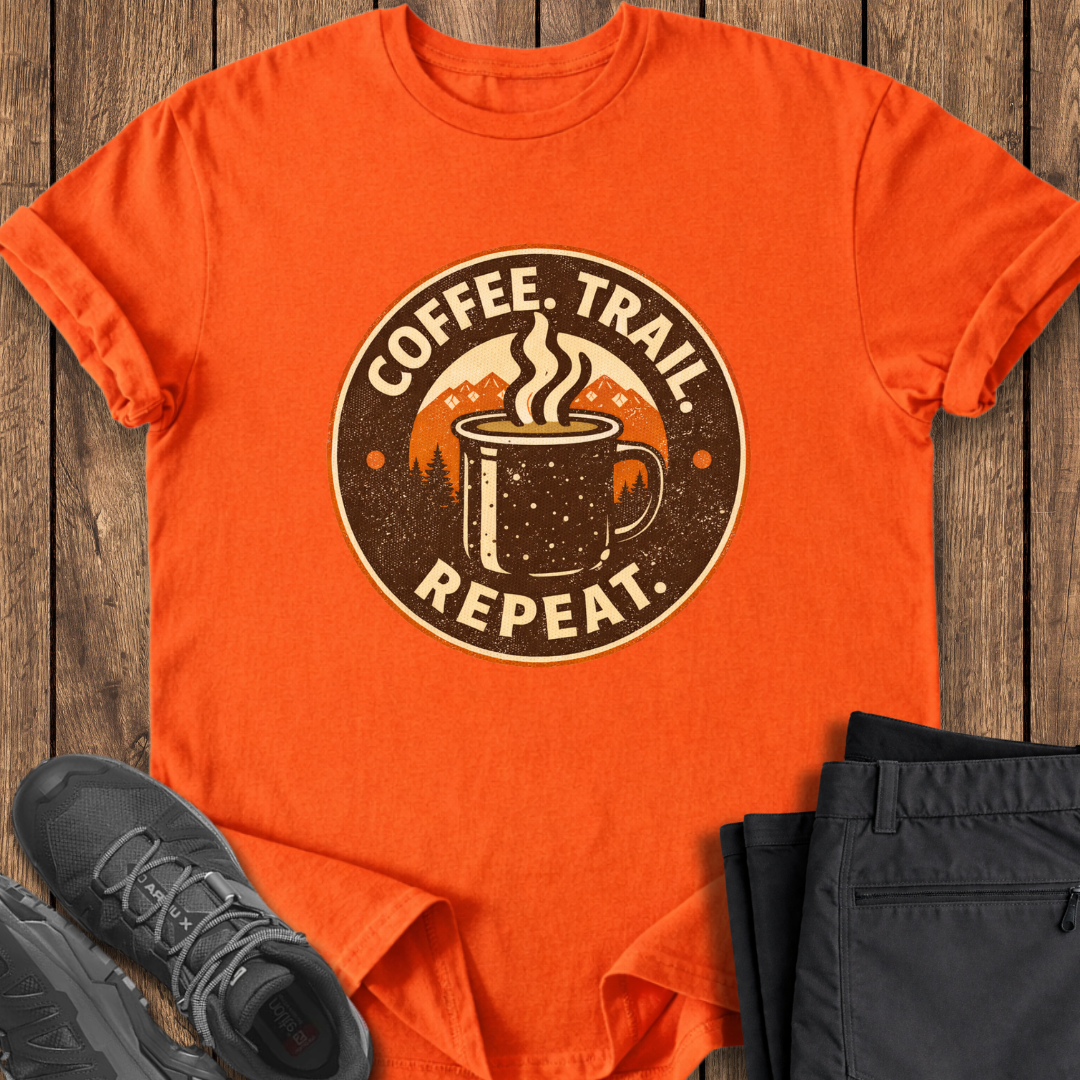Orange t-shirt with 'Coffee. Trail. Repeat.' graphic on a wooden surface with black shoes and pants. coffee hiking t shirt