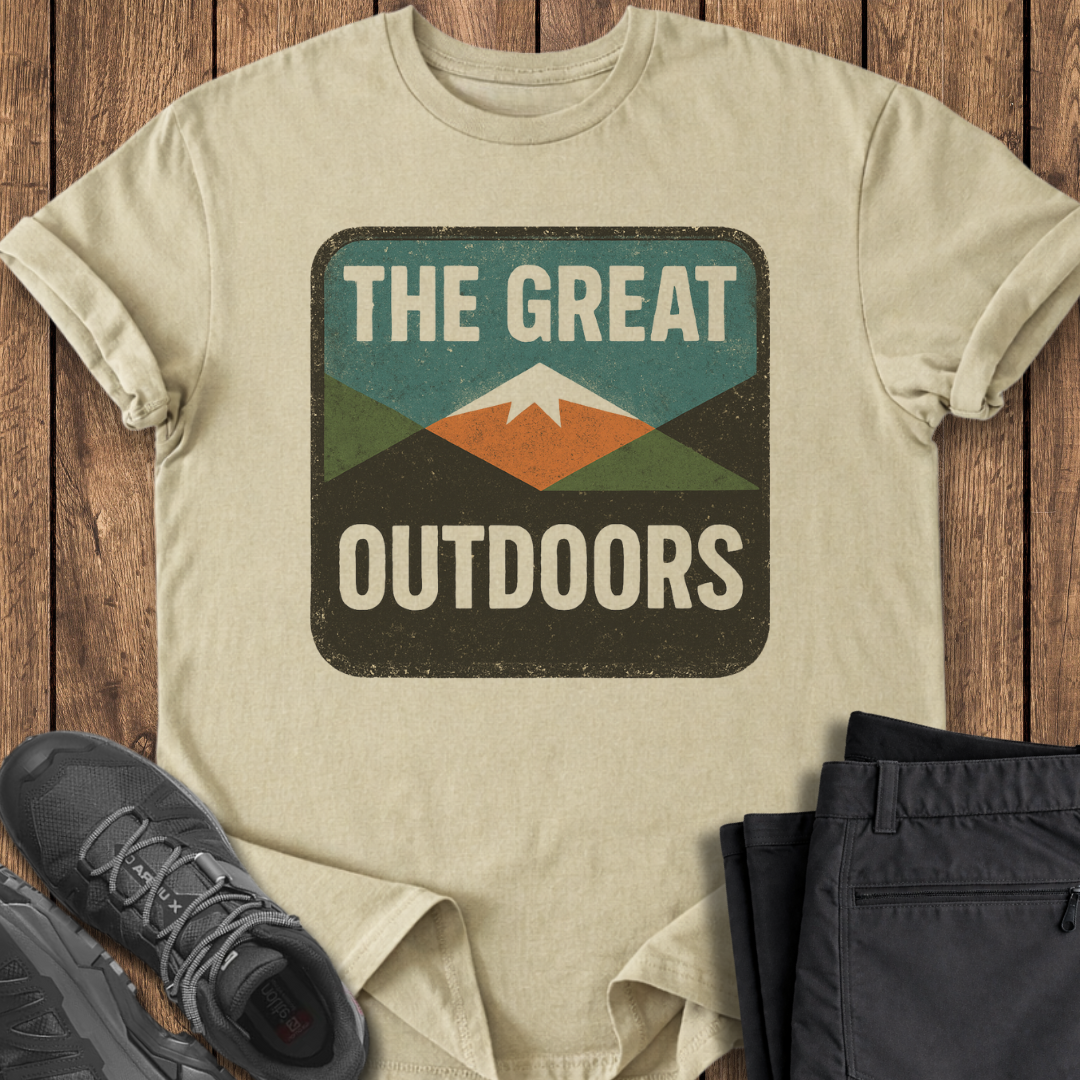 Beige t-shirt with 'The Great Outdoors' graphic on a wooden surface with shoes and pants. outdoor t shirt