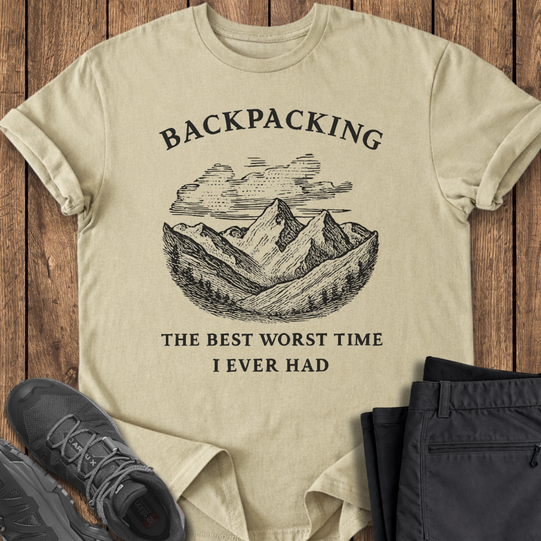 Beige t-shirt with mountain graphic and text on a wooden surface with shoes and pants. funny backpacking t shirt