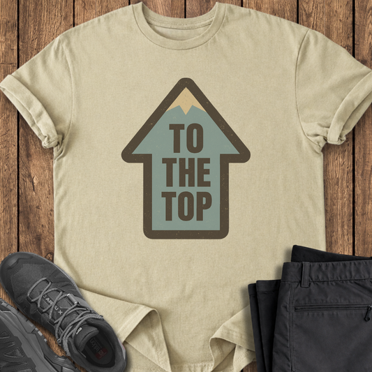 Beige t-shirt with 'To The Top' graphic on a wooden surface with shoes and pants.