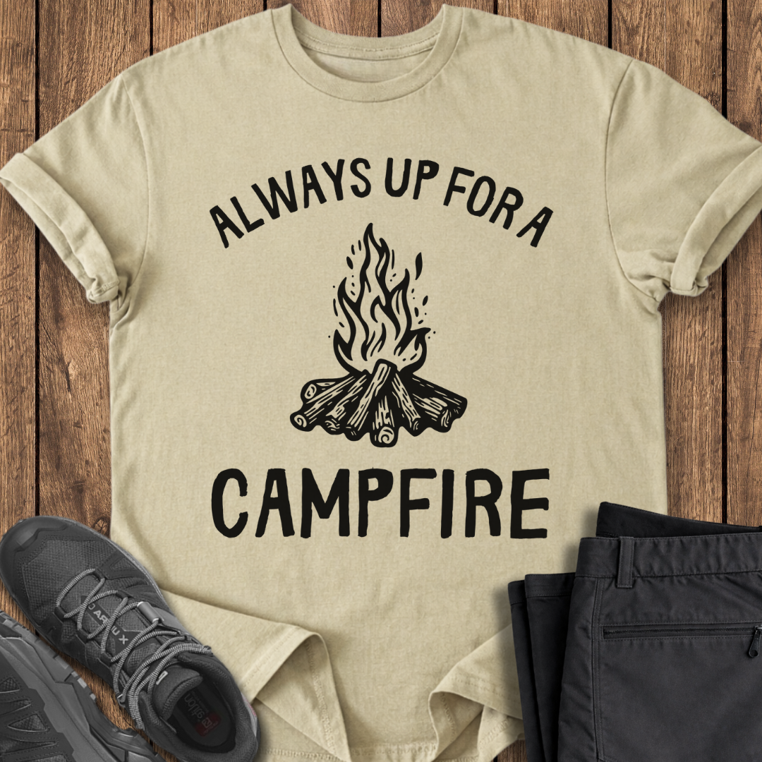 Beige t-shirt with 'Always Up For a Campfire' graphic on a wooden surface with shoes and pants. camping shirt
