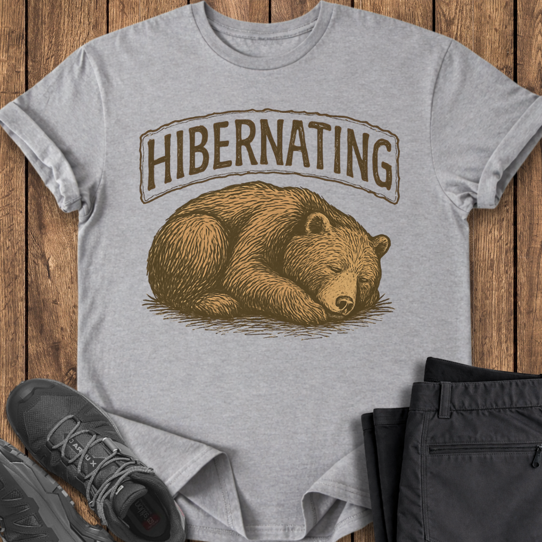 Gray t-shirt with bear graphic and 'Hibernating' text on a wooden surface with shoes and pants.