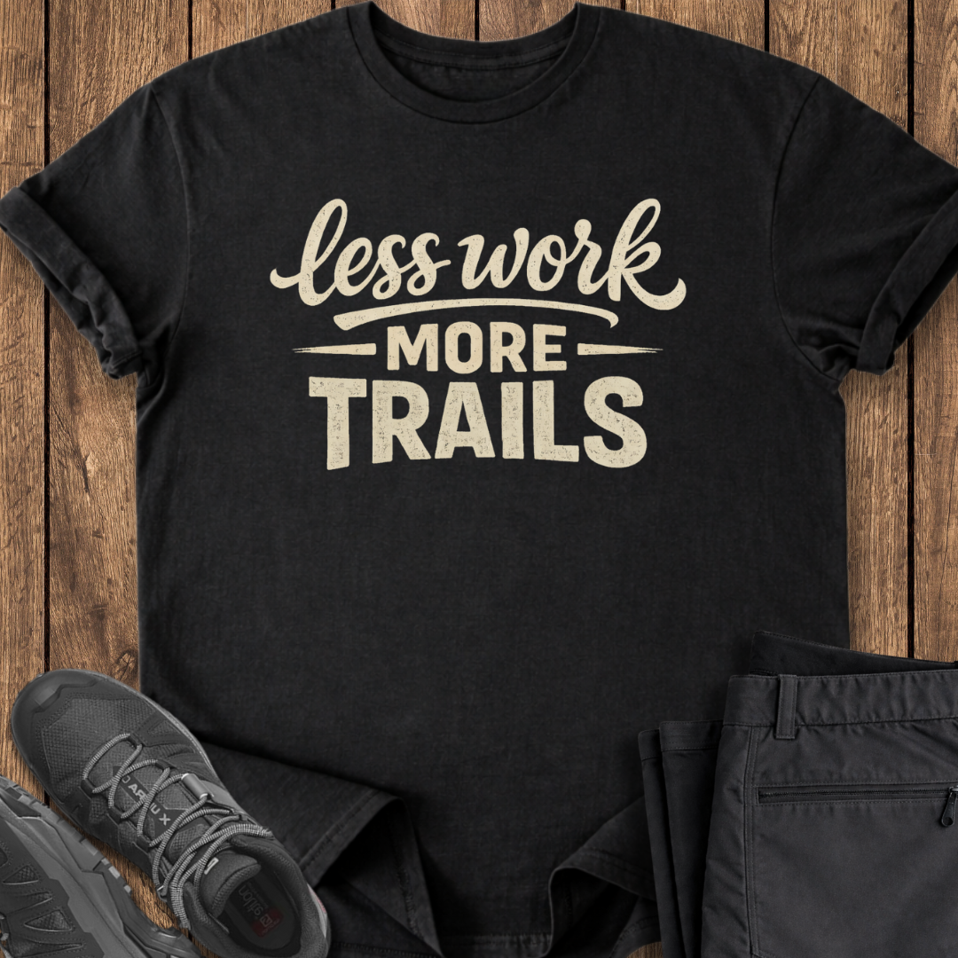 Black t-shirt with 'less work more trails' text on a wooden surface with shoes and pants.