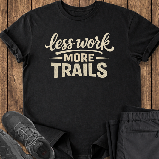 Black t-shirt with 'less work more trails' text on a wooden surface with shoes and pants.