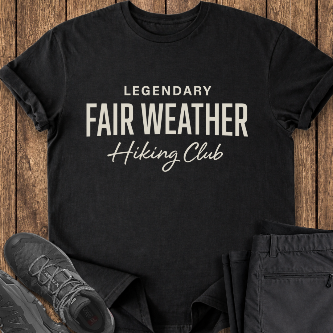 Black t-shirt with 'Legendary Fair Weather Hiking Club' text on a wooden surface with shoes and pants.