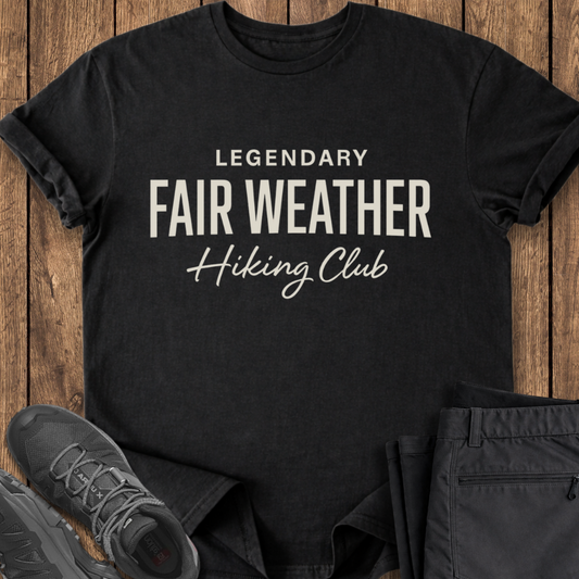 Black t-shirt with 'Legendary Fair Weather Hiking Club' text on a wooden surface with shoes and pants.