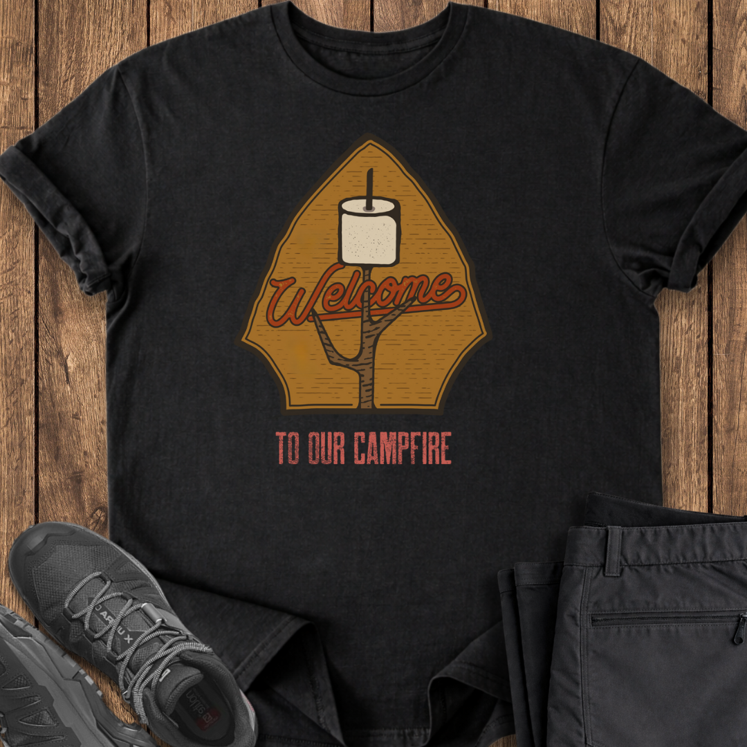 Black t-shirt with 'Welcome to Our Campfire' design on a wooden surface with shoes and pants. camping camp outdoors t shirts