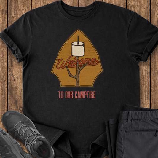 Black t-shirt with 'Welcome to Our Campfire' design on a wooden surface with shoes and pants. camping camp outdoors t shirts