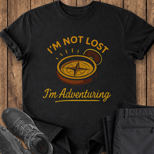 Black t-shirt with 'I'm Not Lost, I'm Adventuring' vintage compass design on a wooden surface with shoes and pants. hiking adventure t shirt