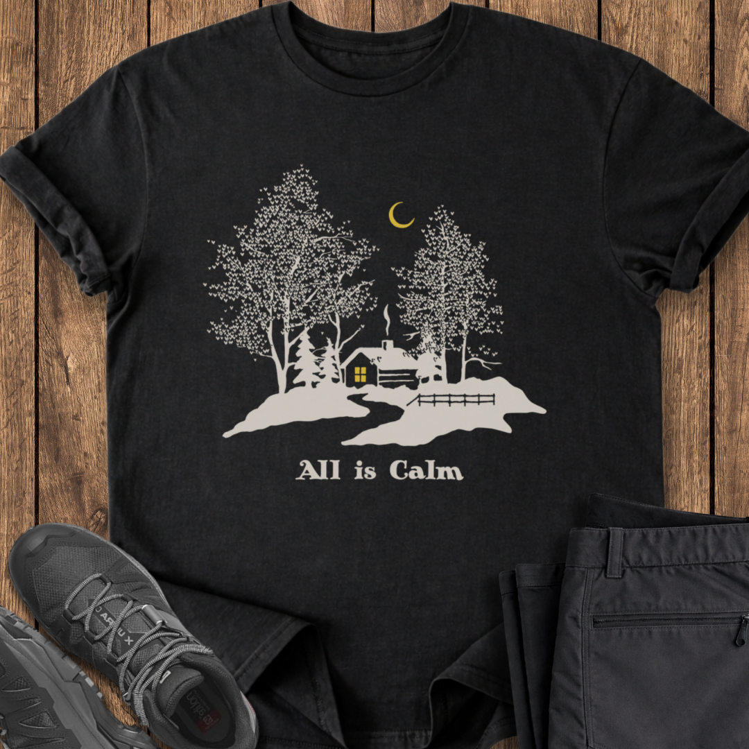 Black t-shirt with a cabin and tree design on a wooden surface with shoes and pants. winter cabin t shirt