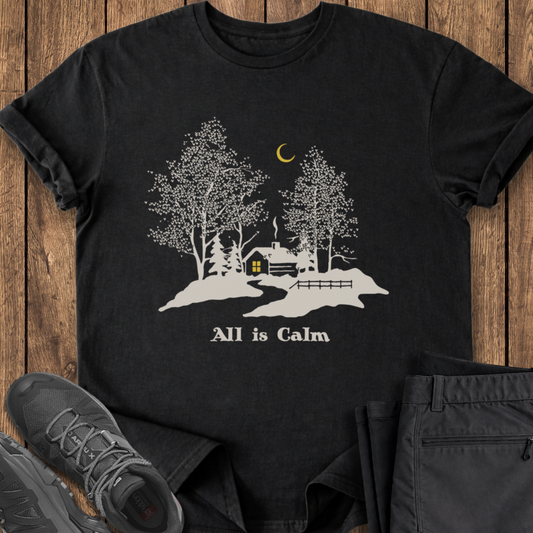 Black t-shirt with a cabin and tree design on a wooden surface with shoes and pants. winter cabin t shirt