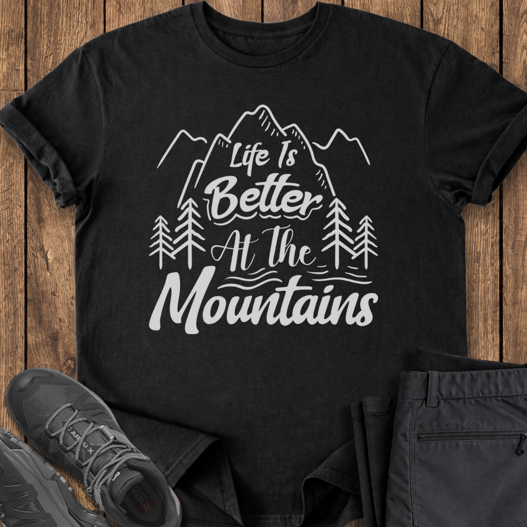 Black t-shirt with 'Life is Better at the Mountains' design on a wooden surface with shoes and pants.