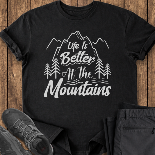 Black t-shirt with 'Life is Better at the Mountains' design on a wooden surface with shoes and pants.