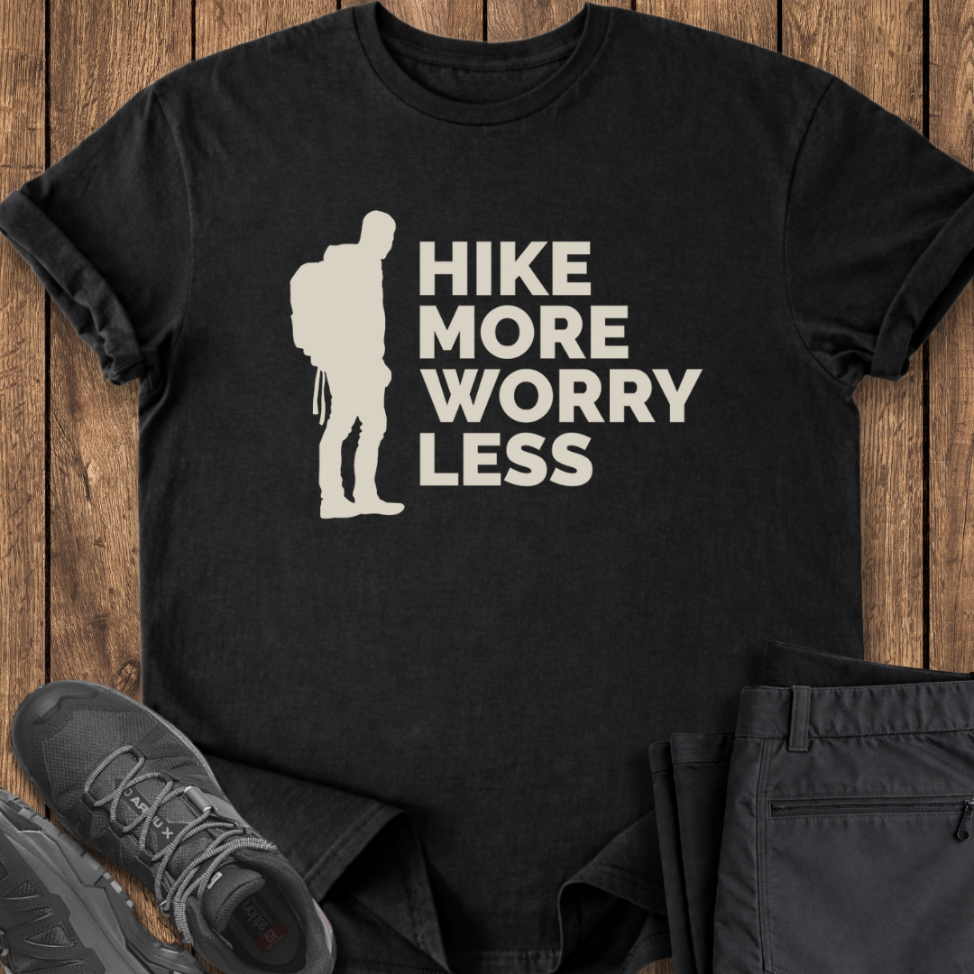 Black t-shirt with 'Hike More Worry Less' design on a wooden surface with hiking shoes and pants.