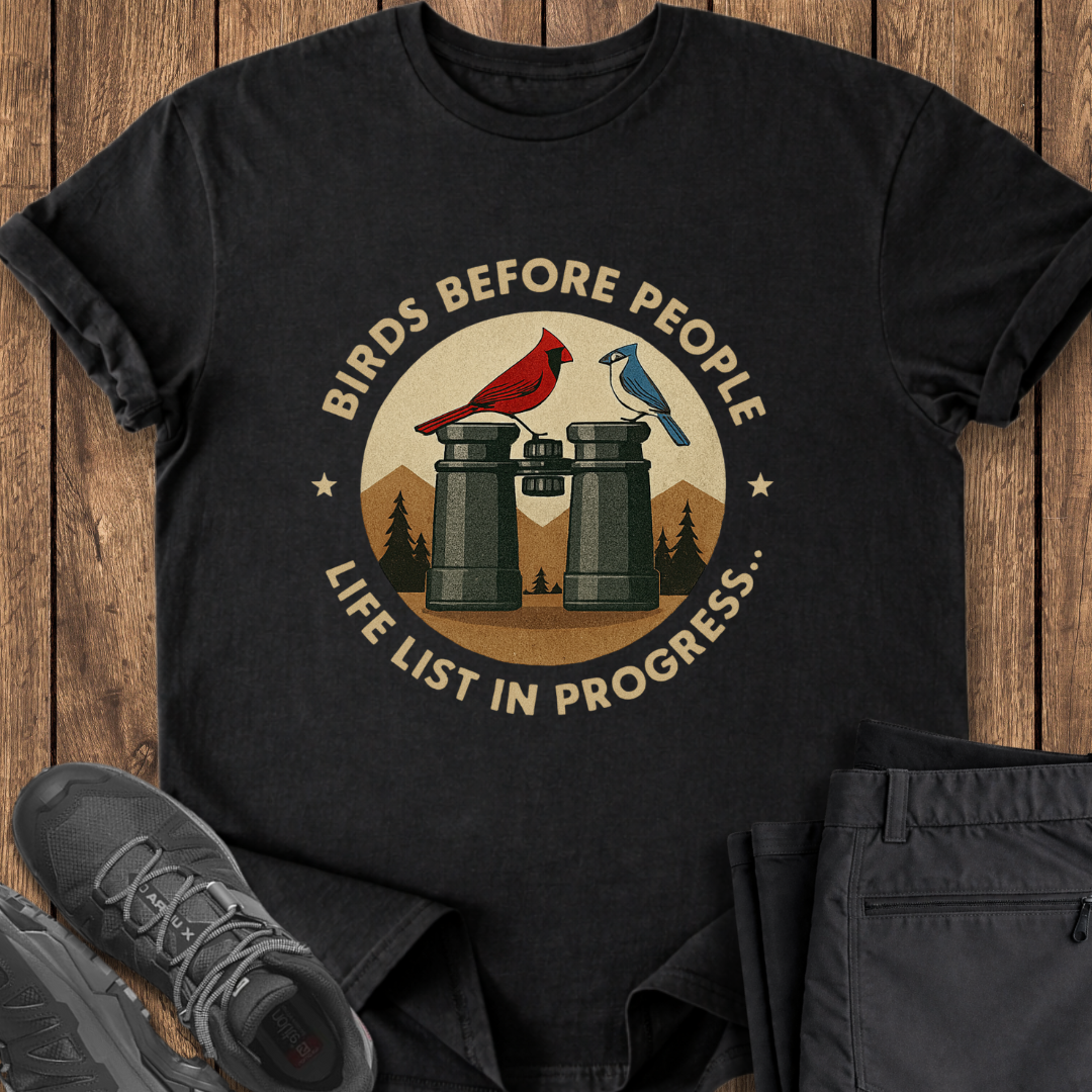 Black t-shirt with 'Birds Before People' graphic design on a wooden surface with shoes and pants.