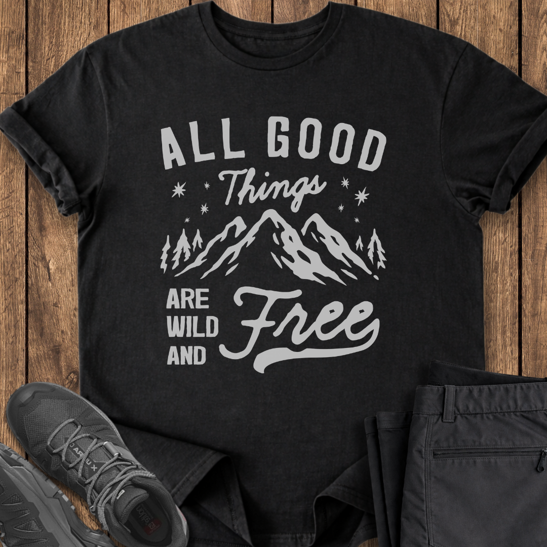 Black t-shirt with 'All Good Things Are Wild and Free' text and mountain graphic on a wooden surface with shoes and pants.