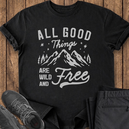 Black t-shirt with 'All Good Things Are Wild and Free' text and mountain graphic on a wooden surface with shoes and pants.