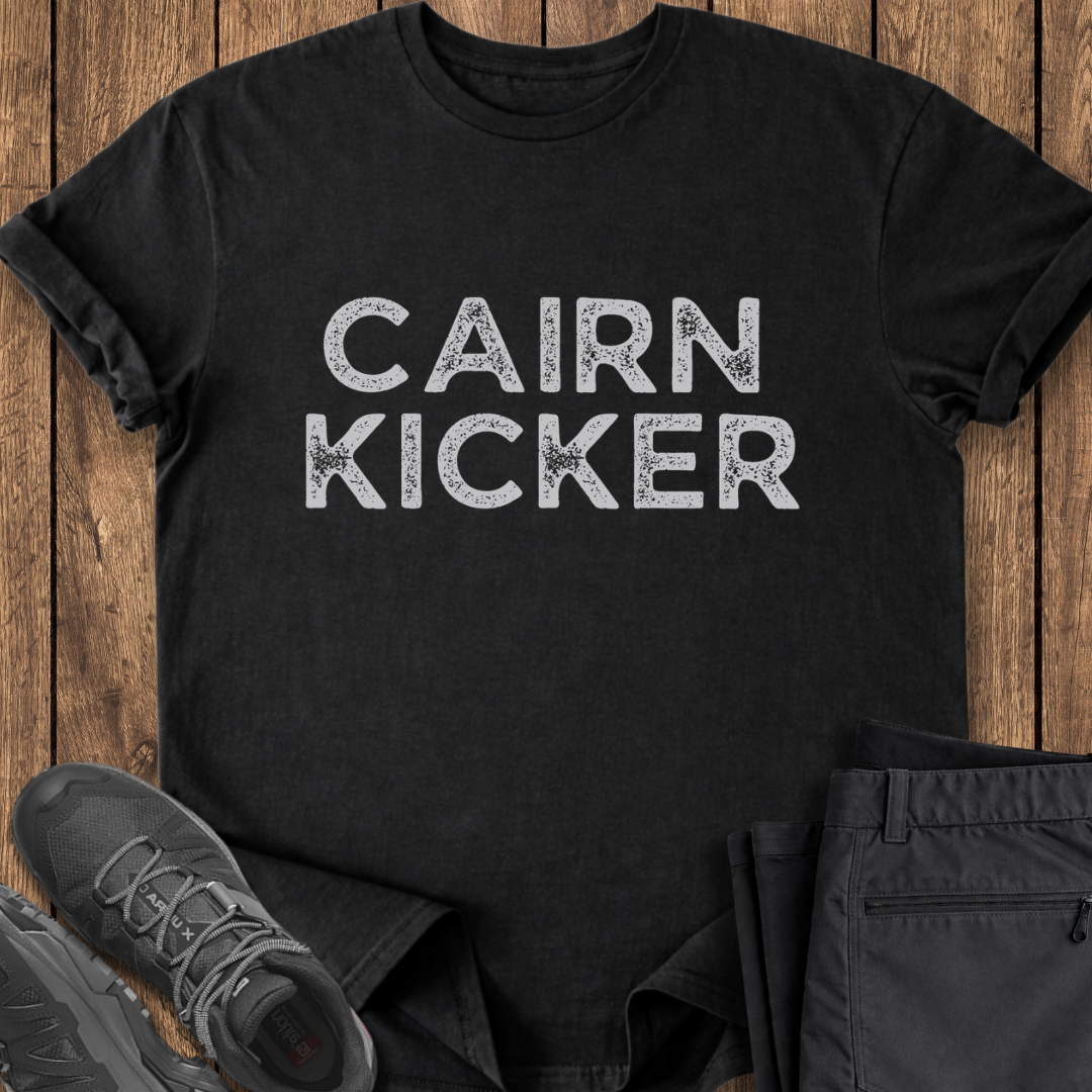 Black t-shirt with 'CAIRN KICKER' text on a wooden surface with shoes and pants. controversial hiking t shirt