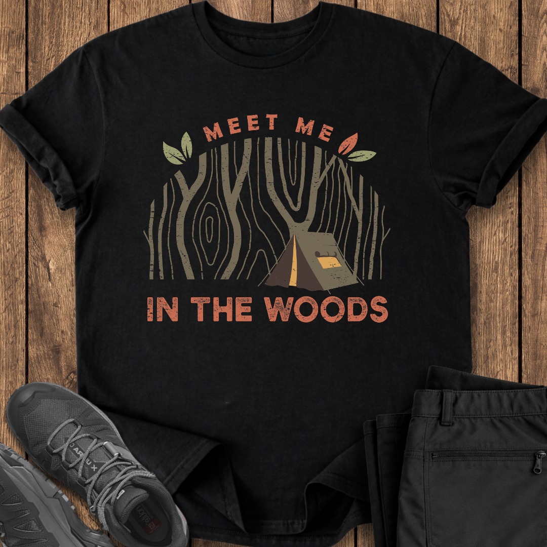 Black t-shirt with 'Meet Me in the Woods' design on a wooden surface with shoes and pants.