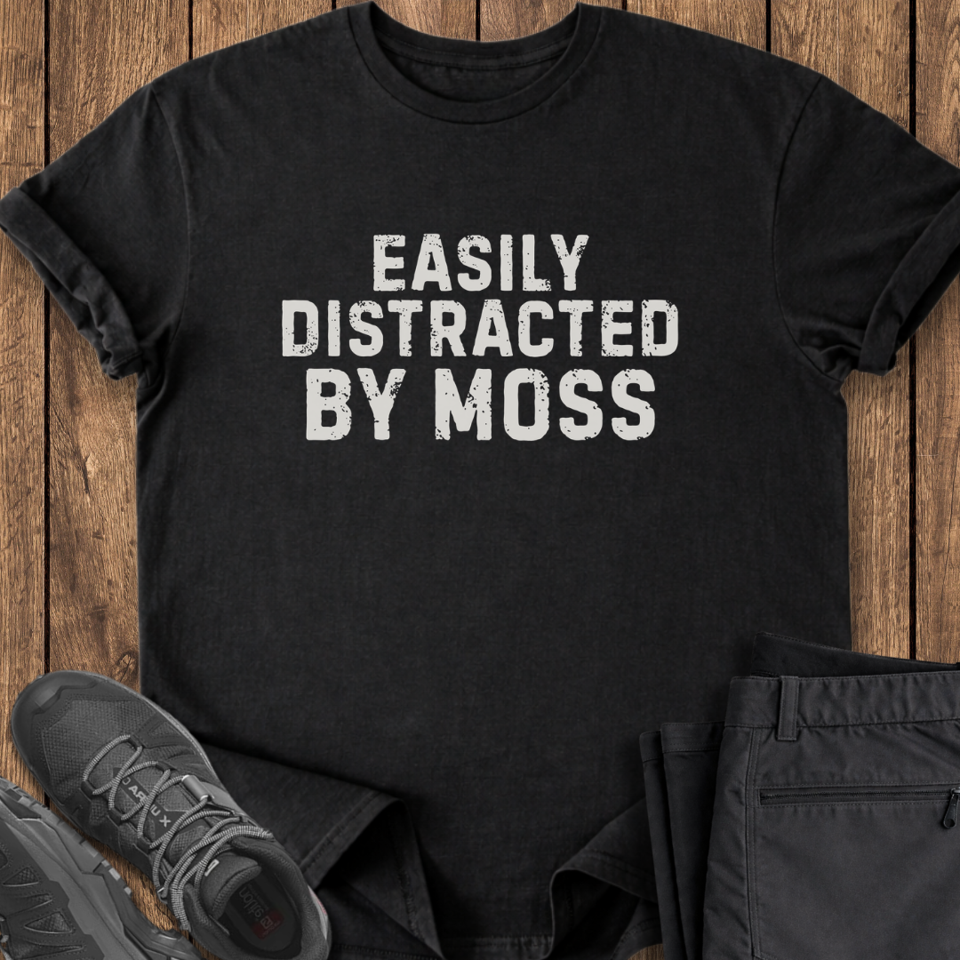 Black t-shirt with 'Easily Distracted By Moss' text on a wooden surface with shoes and pants. funny hiing t shirt