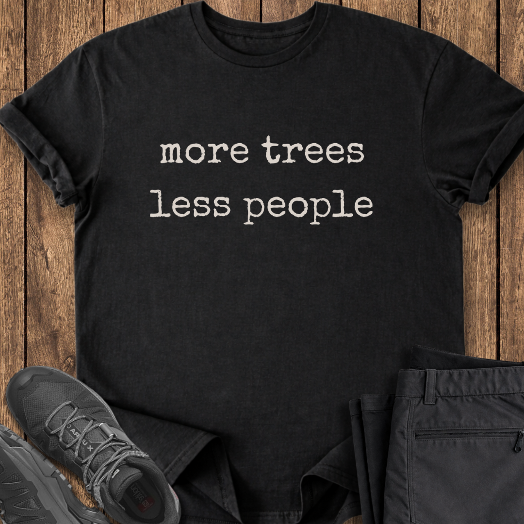 Black t-shirt with 'more trees less people' text on a wooden surface with shoes and pants.