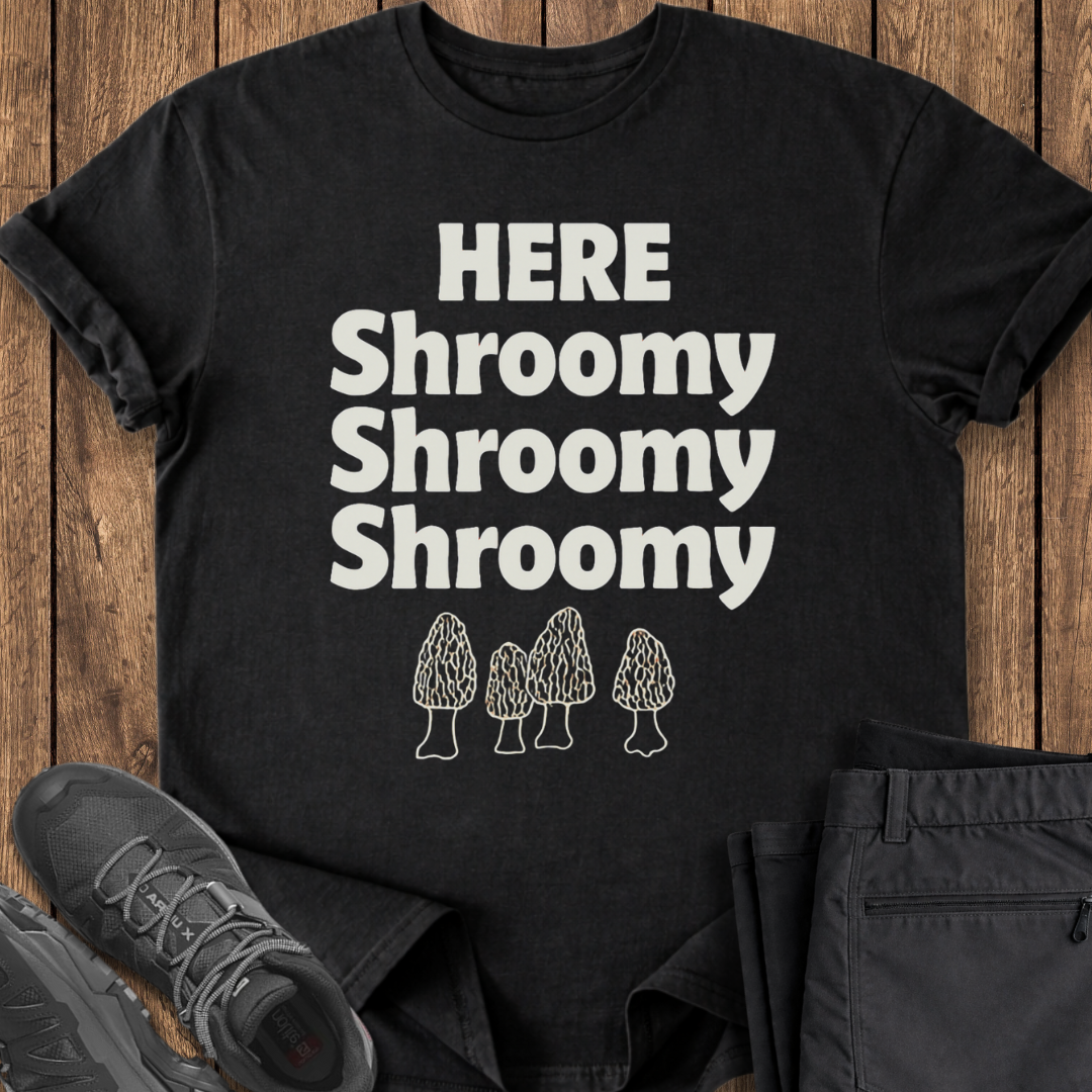 Black t-shirt with 'HERE Shroomy Shroomy Shroomy' text and mushroom graphics on a wooden surface.