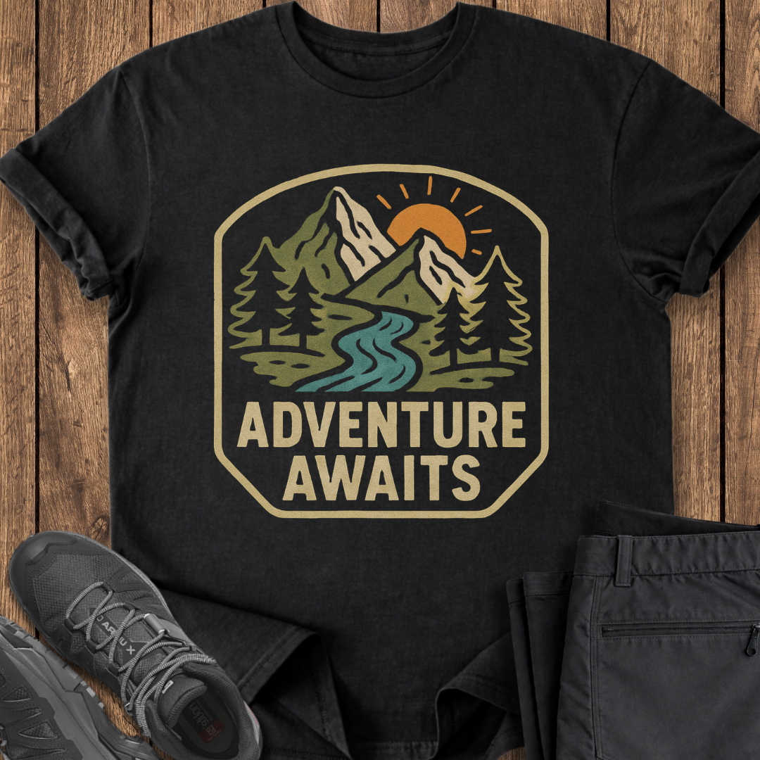Black t-shirt with 'Adventure Awaits' graphic on a wooden surface with shoes and pants. mountain hiking adventure t shirt