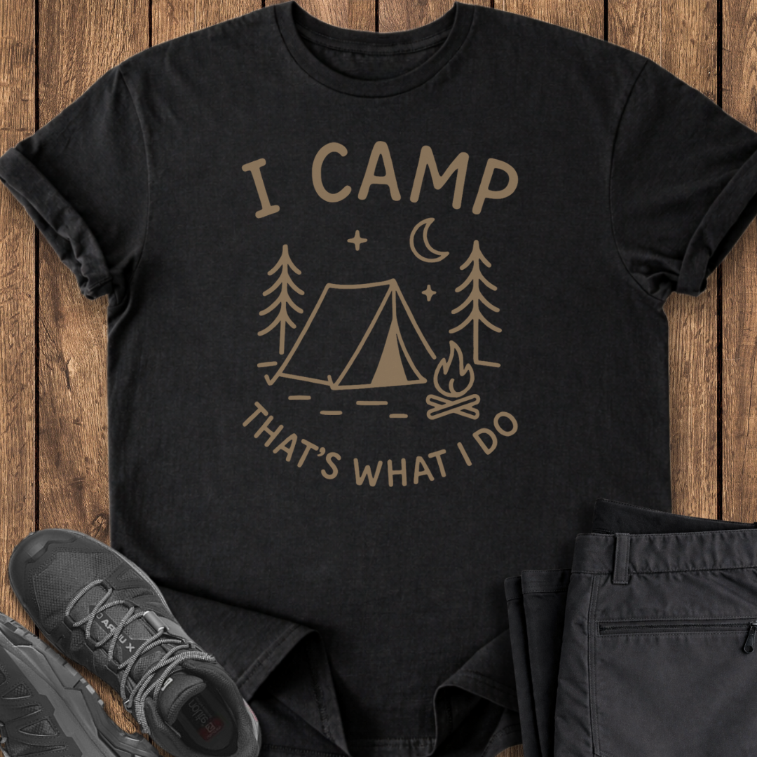 Black t-shirt with 'I Camp, That's What I Do' design on a wooden surface with shoes and pants.